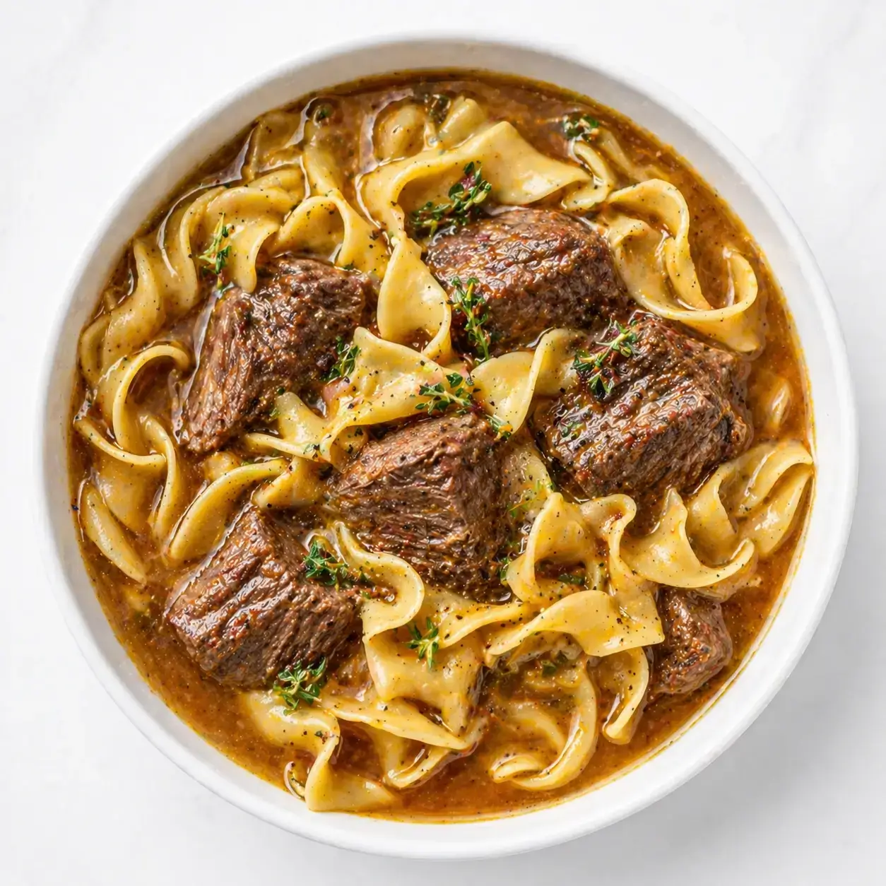 Beef and noodles in a white bowl with tender braised beef chunks, egg noodles, rich brown gravy, fresh thyme, and black pepper overhead view