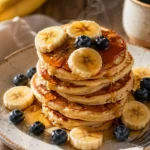 Fluffy mini banana pancakes stacked on a rustic plate with maple syrup, banana slices, blueberries, and coffee in a cozy breakfast setting