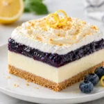 No-bake lemon blueberry dessert bar with graham crust, lemon cheesecake, blueberry topping and whipped cream on plate.