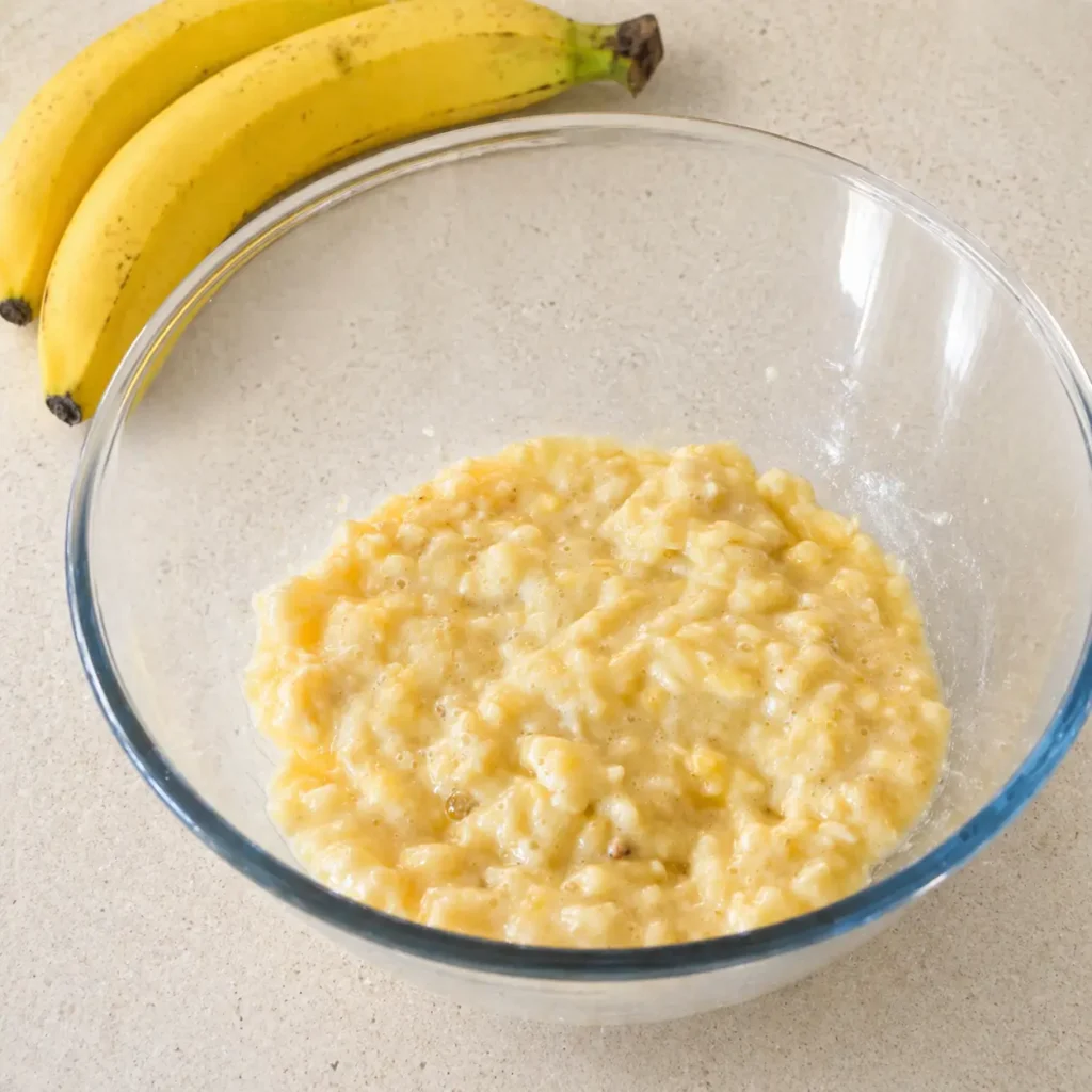 Mashed ripe bananas in a glass mixing bowl with whole bananas on a beige countertop for healthy banana recipe preparation.