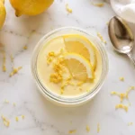 Creamy homemade lemon posset in a glass jar topped with fresh lemon slices and zest on a white marble surface with lemons.