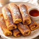 Golden cinnamon sugar French toast roll-ups dusted with powdered sugar and served with warm maple syrup on a white plate.